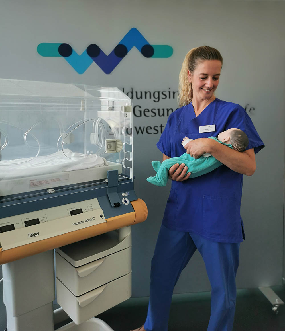 Eine Auszubildende zur Pflegefachfrau im blauen Kittel übt im SkillsLab und holt eine Babypuppe aus einem Inkubator