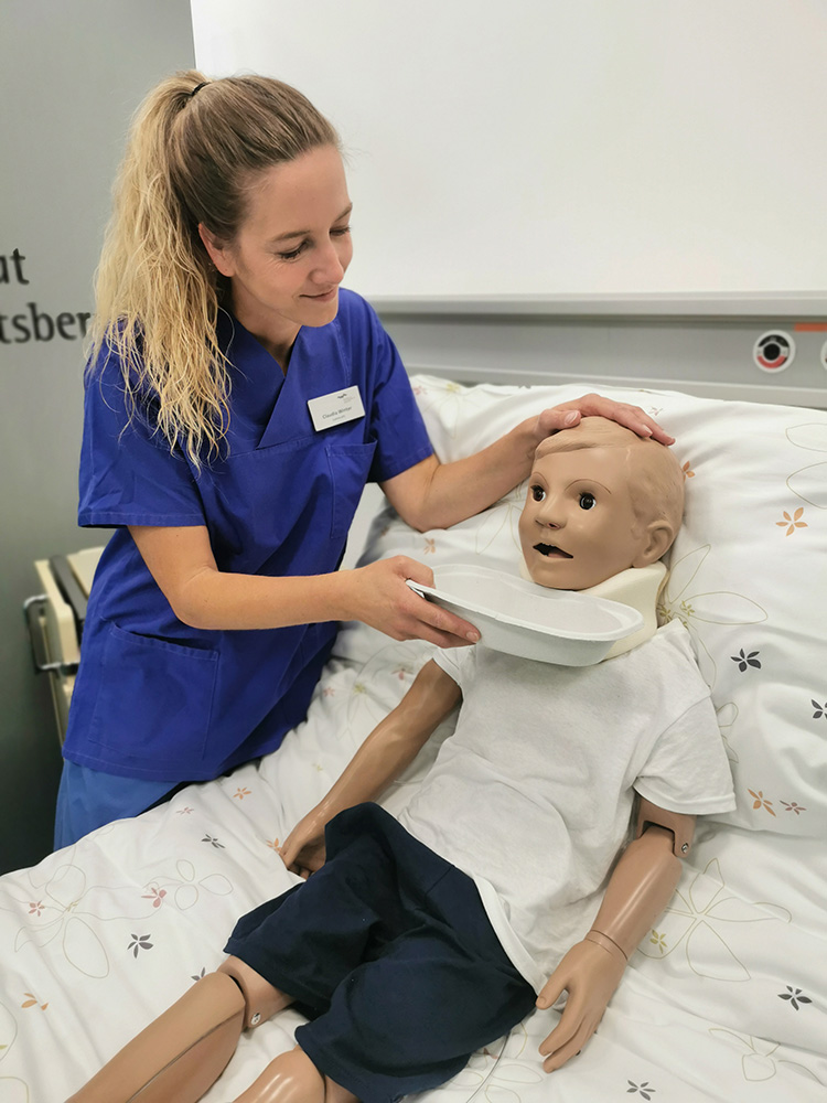 Eine Auszubildende zur Pflegefachfrau im blauen Kittel hält im SkillsLab einer im Bett sitzenden Kinderpuppe eine Auffangschüssel unterhalb des Kopfes