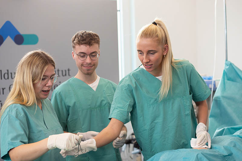 Drei Auszubildende in grünen Kitteln simulieren eine Bauchoperation an einer Puppe im SkillsLab