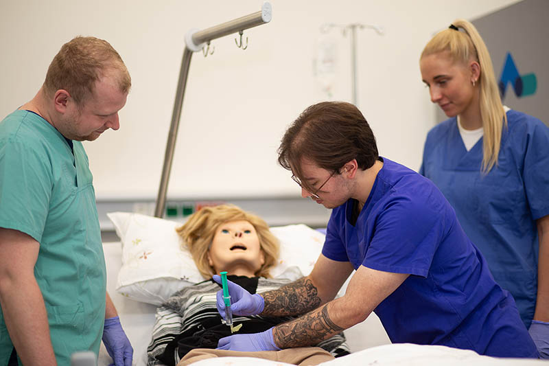 Drei Auszubildende in grünen Kitteln simulieren eine Bauchoperation an einer Puppe im SkillsLab
