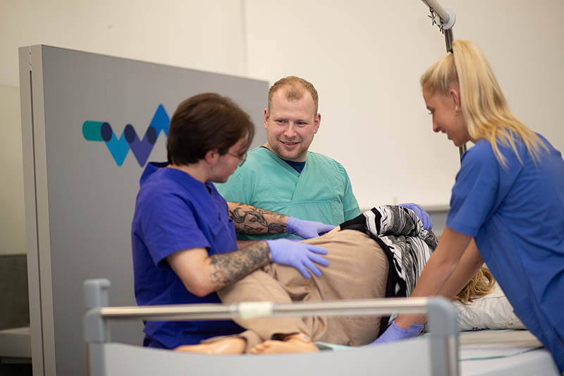 Drei Auszubildende in Kitteln führen eine Behandlung an einer Puppe im SkillsLab durch