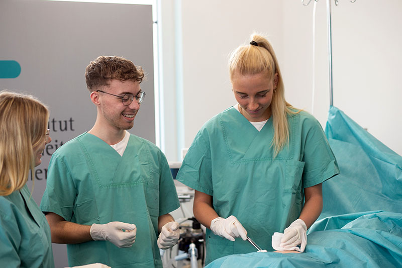 Drei Auszubildende in grünen Kitteln simulieren eine Bauchoperation an einer Puppe im SkillsLab