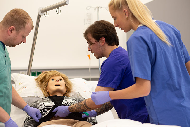 Drei Auszubildende in grünen Kitteln simulieren eine Bauchoperation an einer Puppe im SkillsLab