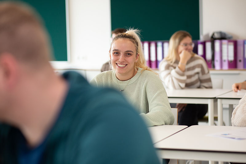 Eine Auszubildende in Freizeitkleidung sitzt an einem Tisch im Klassenraum und lächelt in die Kamera, während andere Schüler im Hintergrund sprechen